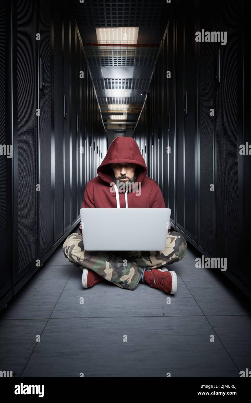 L'hacker in una cappa siede con il computer tra i server, lo scassinatore. Archiviazione dei dati, servizi cloud, interruzione del sistema, attac Foto Stock