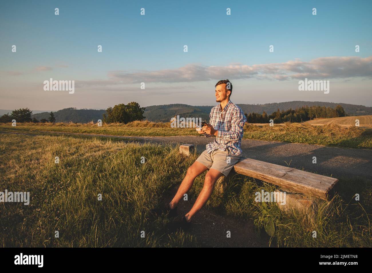 Il fotografo in una maglietta seduta sulla cima di una collina aziona il suo drone per scattare foto del paesaggio sottostante. Progressi tecnologici nelle riprese video. Foto Stock