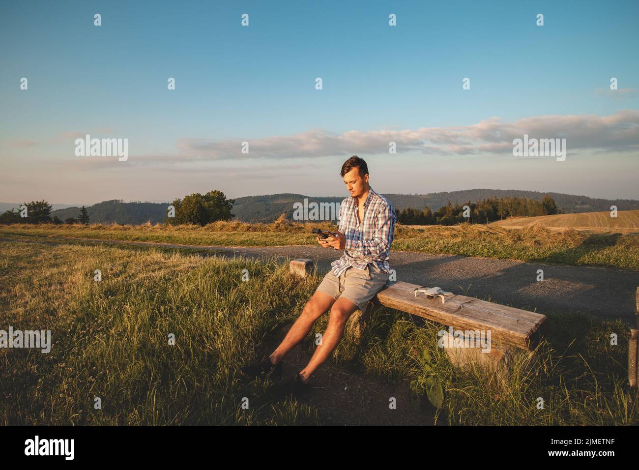 Il fotografo in una maglietta seduta sulla cima di una collina aziona il suo drone per scattare foto del paesaggio sottostante. Progressi tecnologici nelle riprese video. Foto Stock