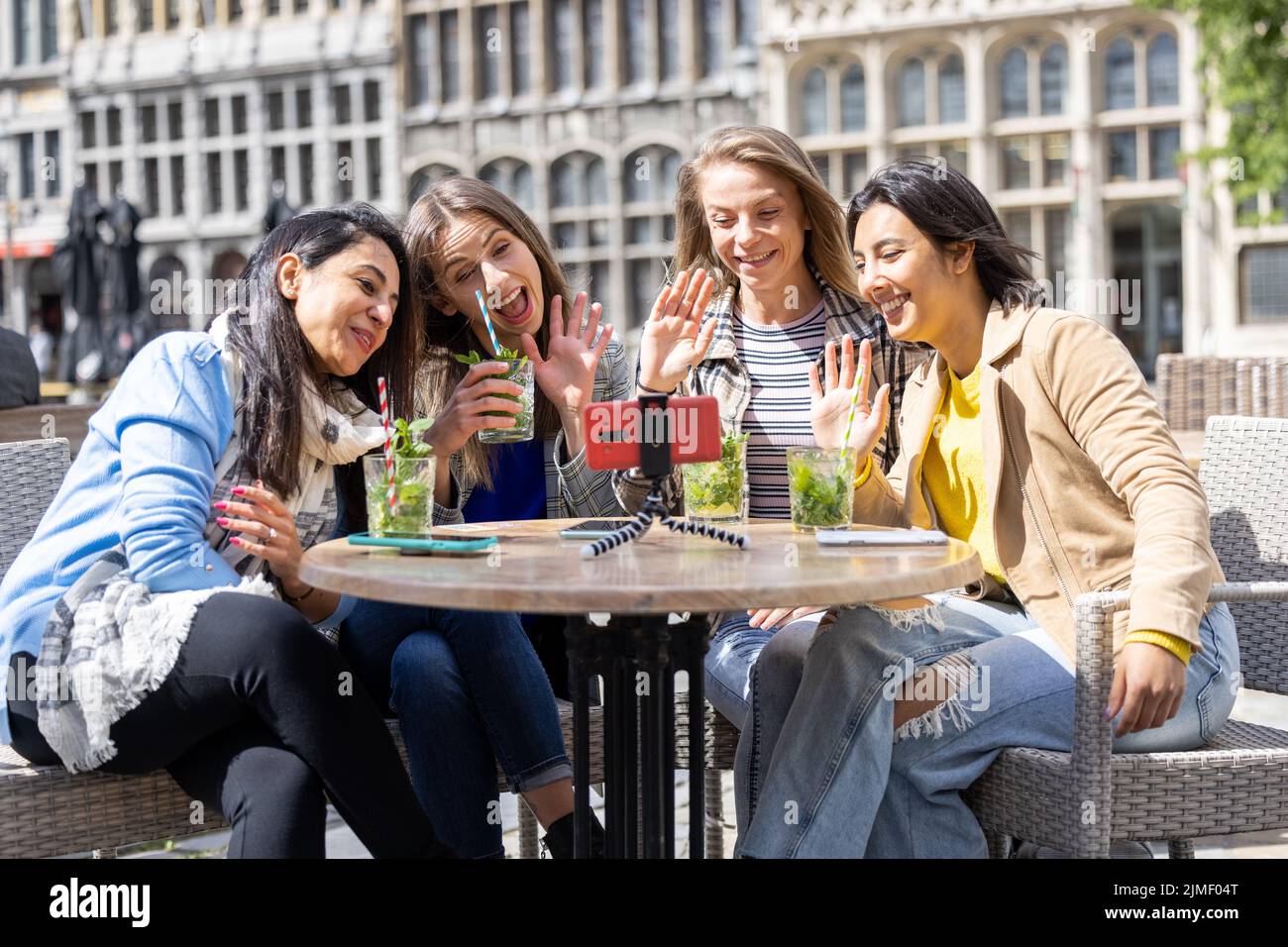 Quattro amici multietnici, turisti o studenti seduti in una caffetteria sul terrazzo del centro della città utilizzando il telefono cellulare per un vid Foto Stock