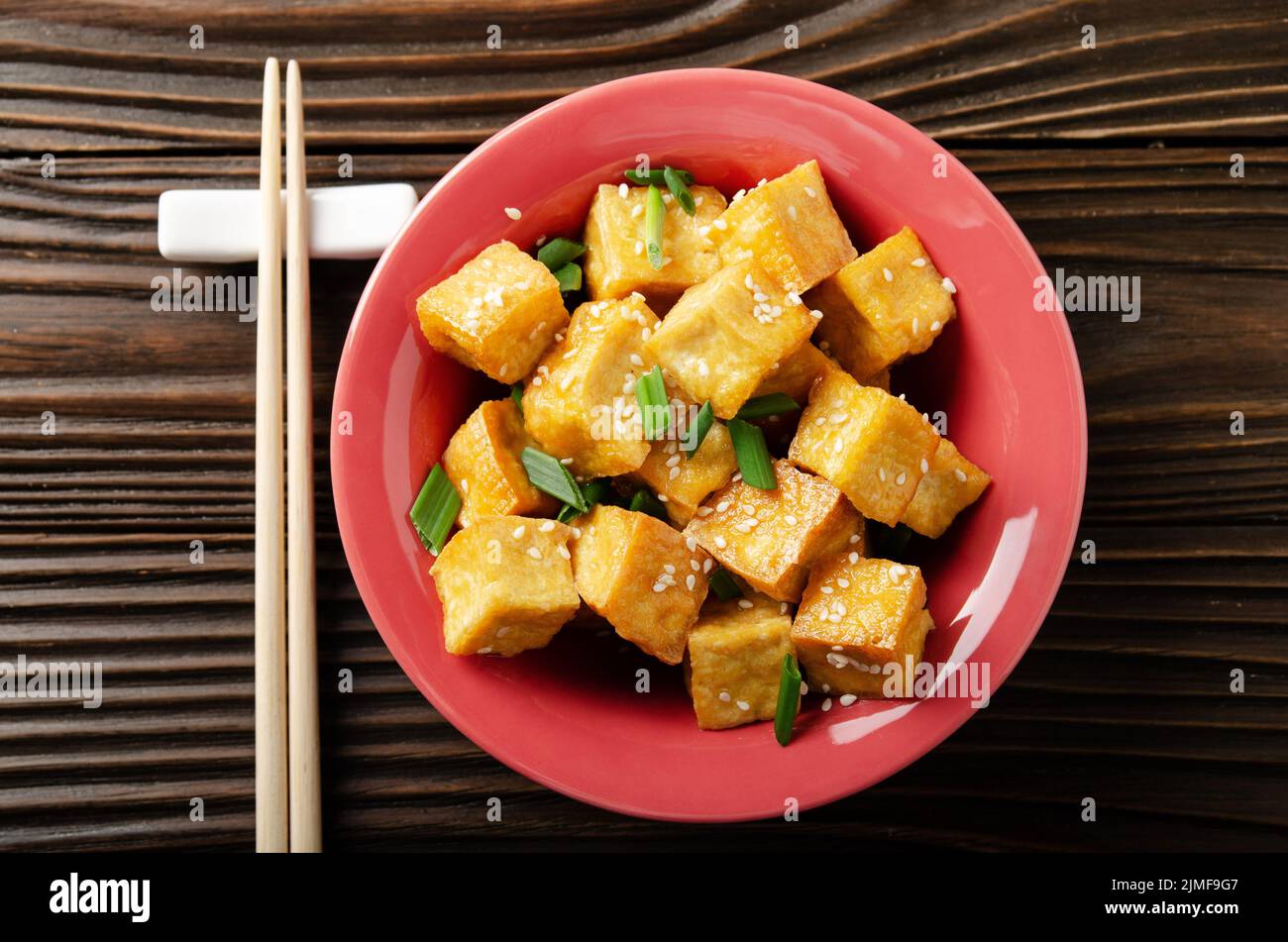 Vista piana a spiovente e profonda mescolare cubetti di tofu fritti con erba cipollina in piatto di argilla su tavolo da cucina in legno Foto Stock