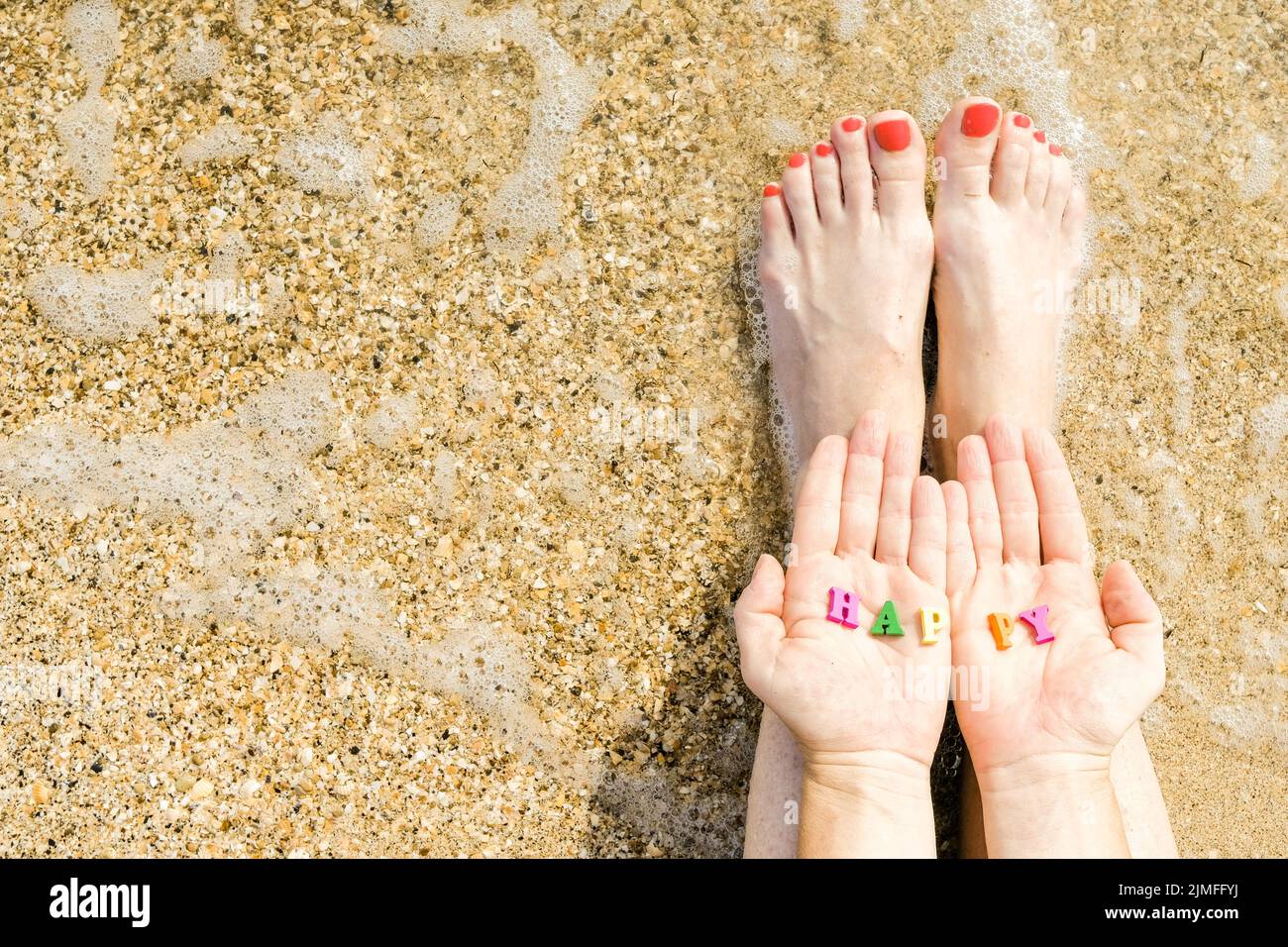 Mani e piedi da donna con pedicure rosse sullo sfondo di sabbia marina e surf. Nelle palme delle lettere con le ins Foto Stock