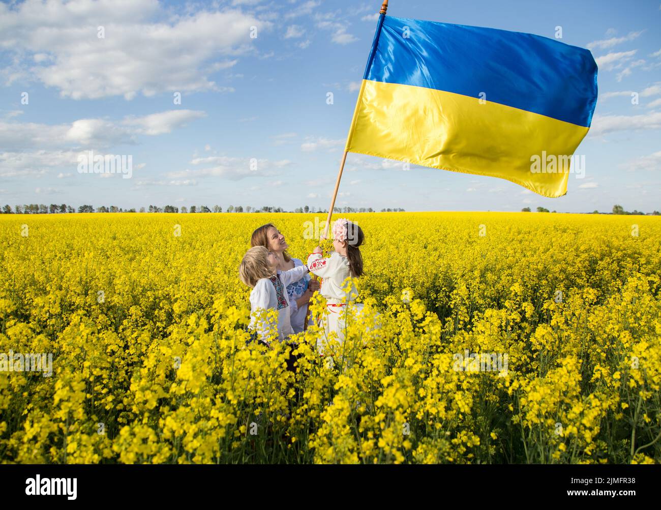 Famiglia ucraina madre, figlio e figlia in camicie ricamate stand tra campo di colza giallo in fiore con grande bandiera di Ucraina. Giorno di sole. Unità Foto Stock