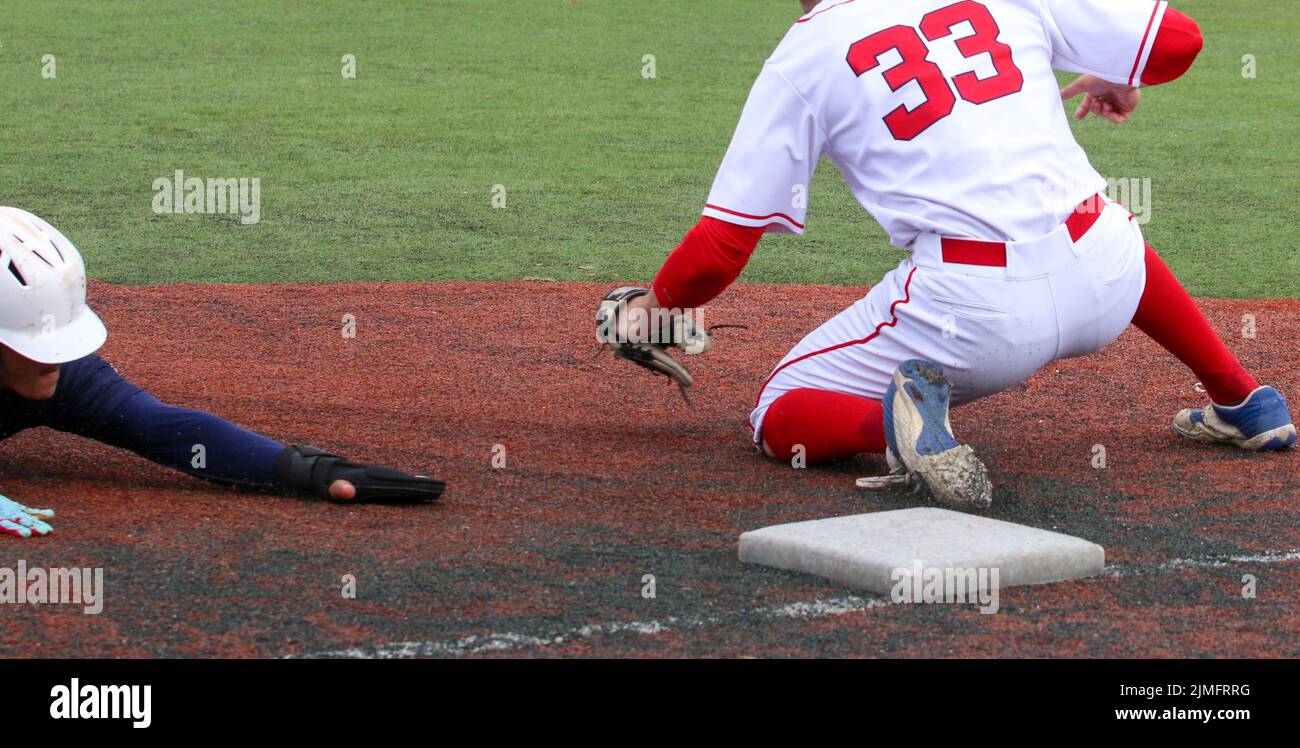 La testa del giocatore di baseball prima scivola nel terzo indossare guanti scorrevoli come il terzo baseman sta cercando di applicare il tag. Foto Stock