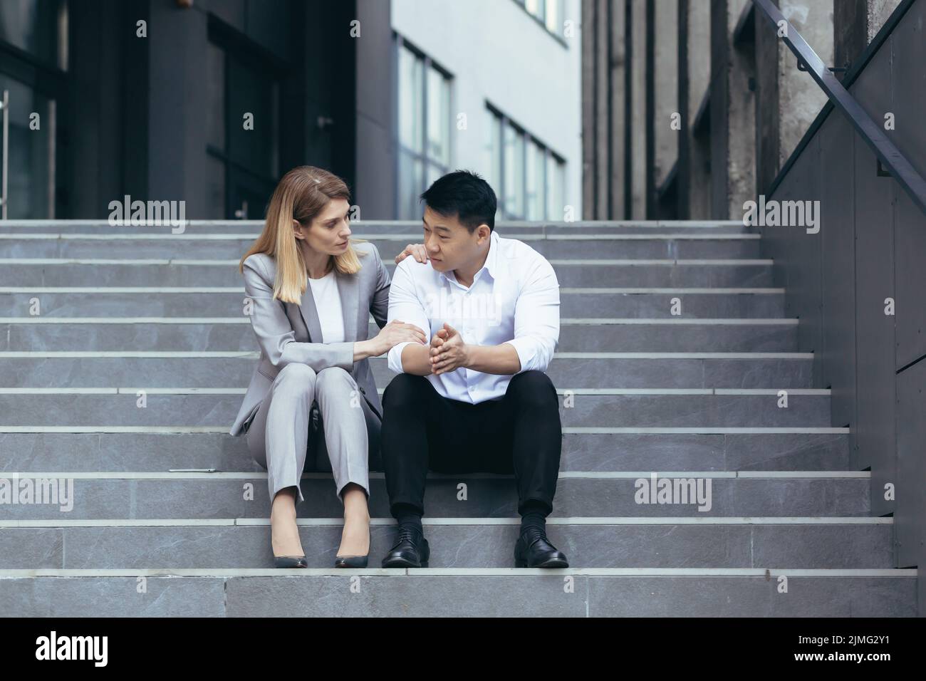 Donna che si prende cura di deluso uomo d'affari asiatico con fallimento di lavoro seduto sulle scale nel centro della città, il sostegno amichevole da parte del collega Foto Stock