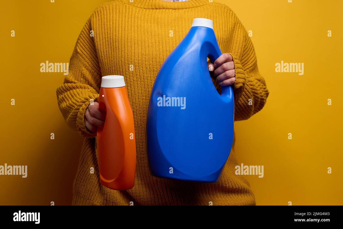 Donna in maglia maglione contiene bottiglie di plastica arancione e blu con gel liquido di lavaggio. Lavanderia e fore, backgro giallo Foto Stock