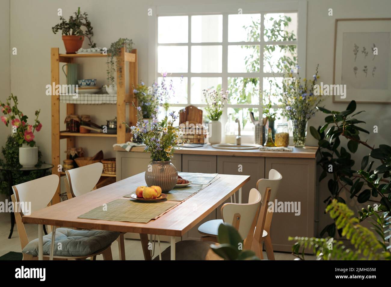 Accogliente cucina con tavolo in legno al centro circondato da lavandino con utensili da cucina e fiori di campo da finestra e piante domestiche verdi Foto Stock