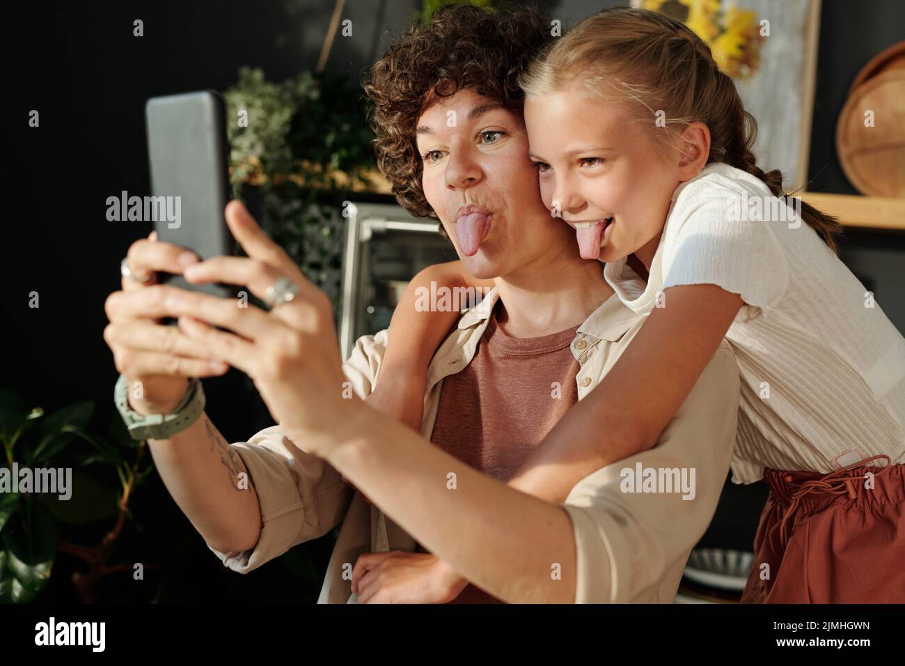 Ragazza giovane carina e sua madre con smartphone che mostra lingue mentre si fa selfie o comunicare in chat video a casa Foto Stock