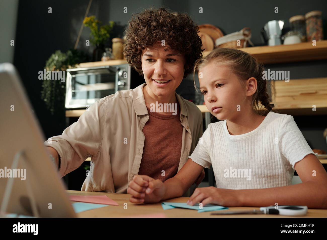 Giovane donna bruna che punta allo schermo del tablet mentre si siede da tavolo accanto a sua figlia e la aiuta con origami a piacere Foto Stock