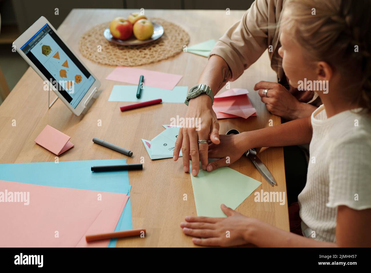 Giovane donna e sua figlia imparando a fare origami a piacere mentre si siede da tavolo e guardando video corso on-line o formazione Foto Stock