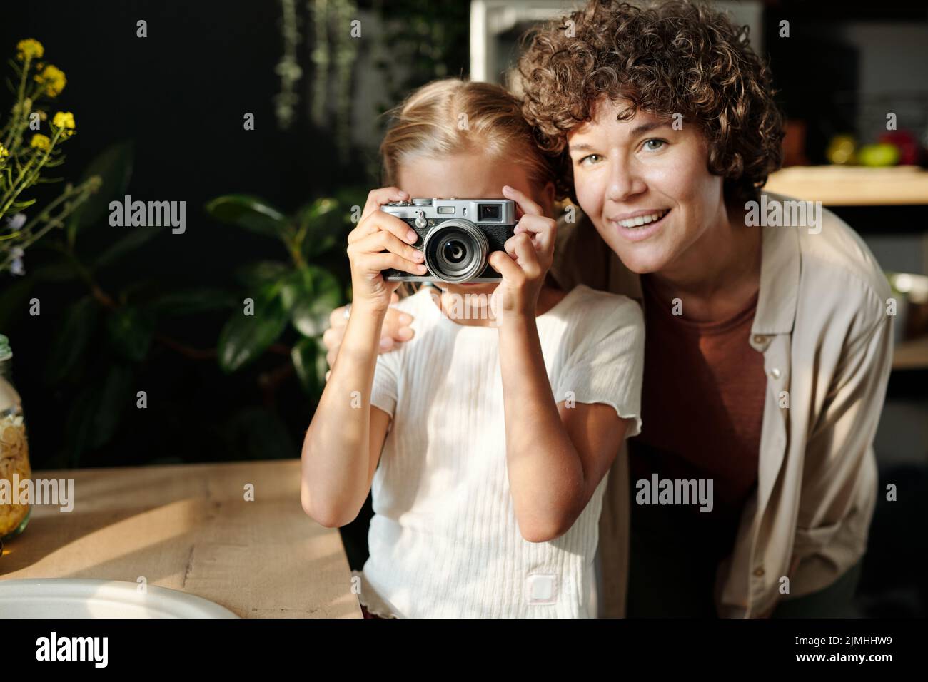 Giovane donna allegra che guarda la macchina fotografica mentre si alza accanto a sua figlia giovane scattando foto su fotocamera a casa Foto Stock