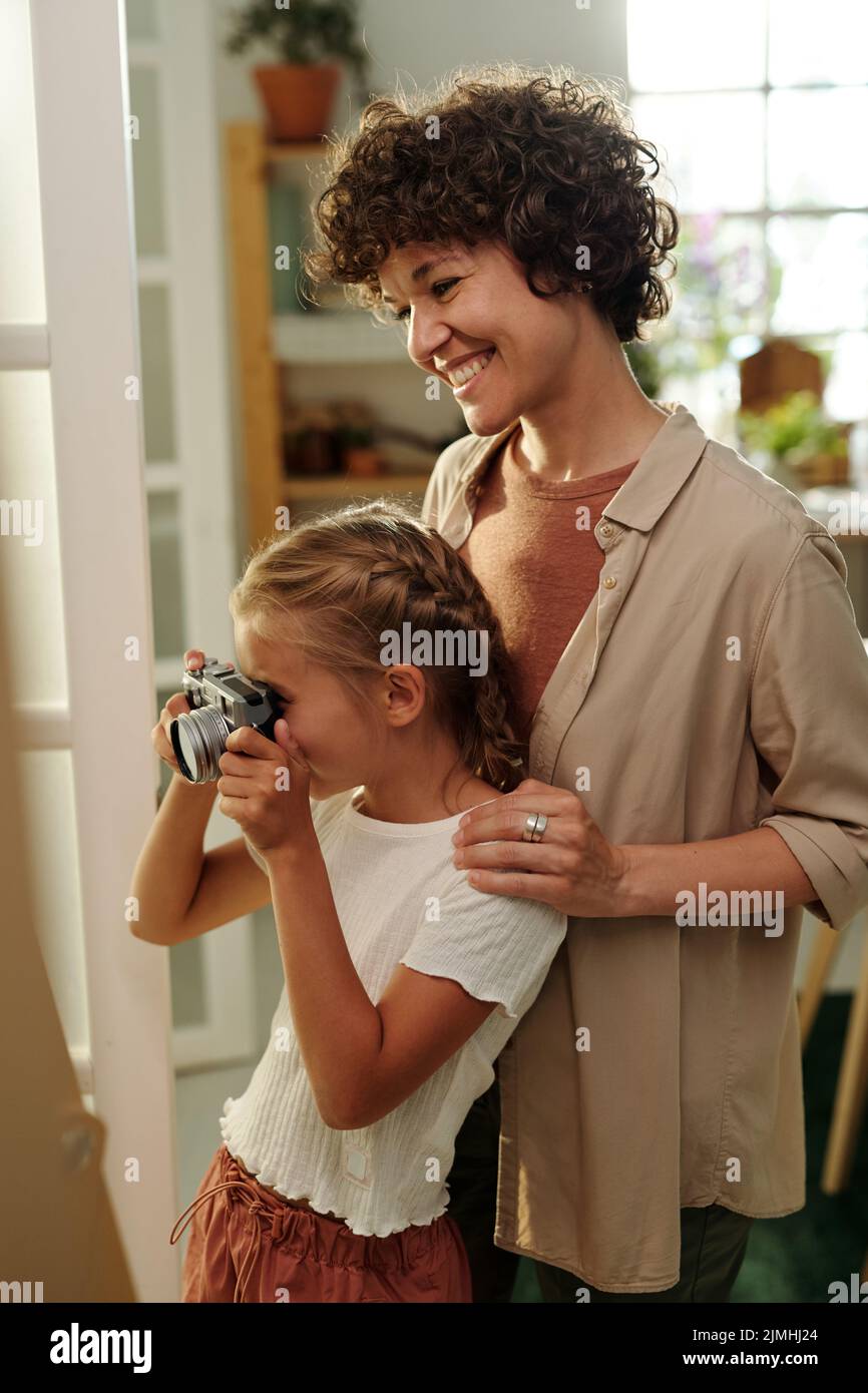 Giovane donna allegra in casualwear in piedi accanto a sua figlia giovane con retro fotocamera sparare lei e madre Foto Stock