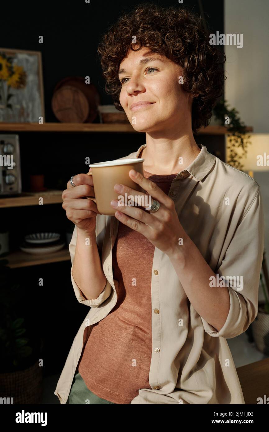 Giovane bruna sorridente con capelli corti ricci scuri che tengono la tazza con il tè o il caffè mentre si alza davanti alla finestra al mattino Foto Stock