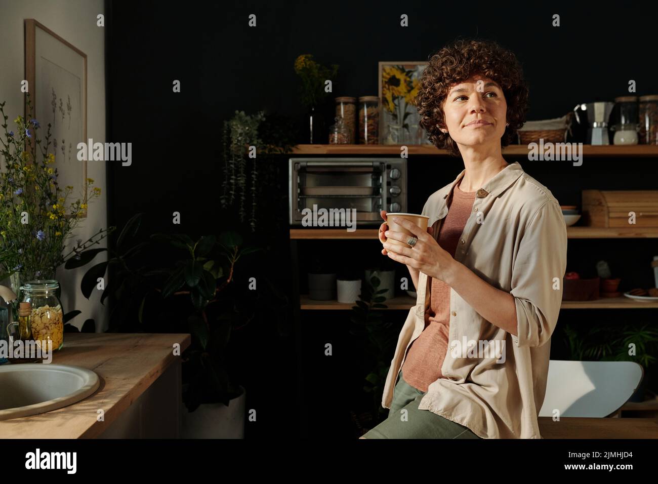 Giovane donna mindful bruna in casualwear che tiene una tazza di tè o caffè mentre si siede sul tavolo da cucina e si riposa Foto Stock