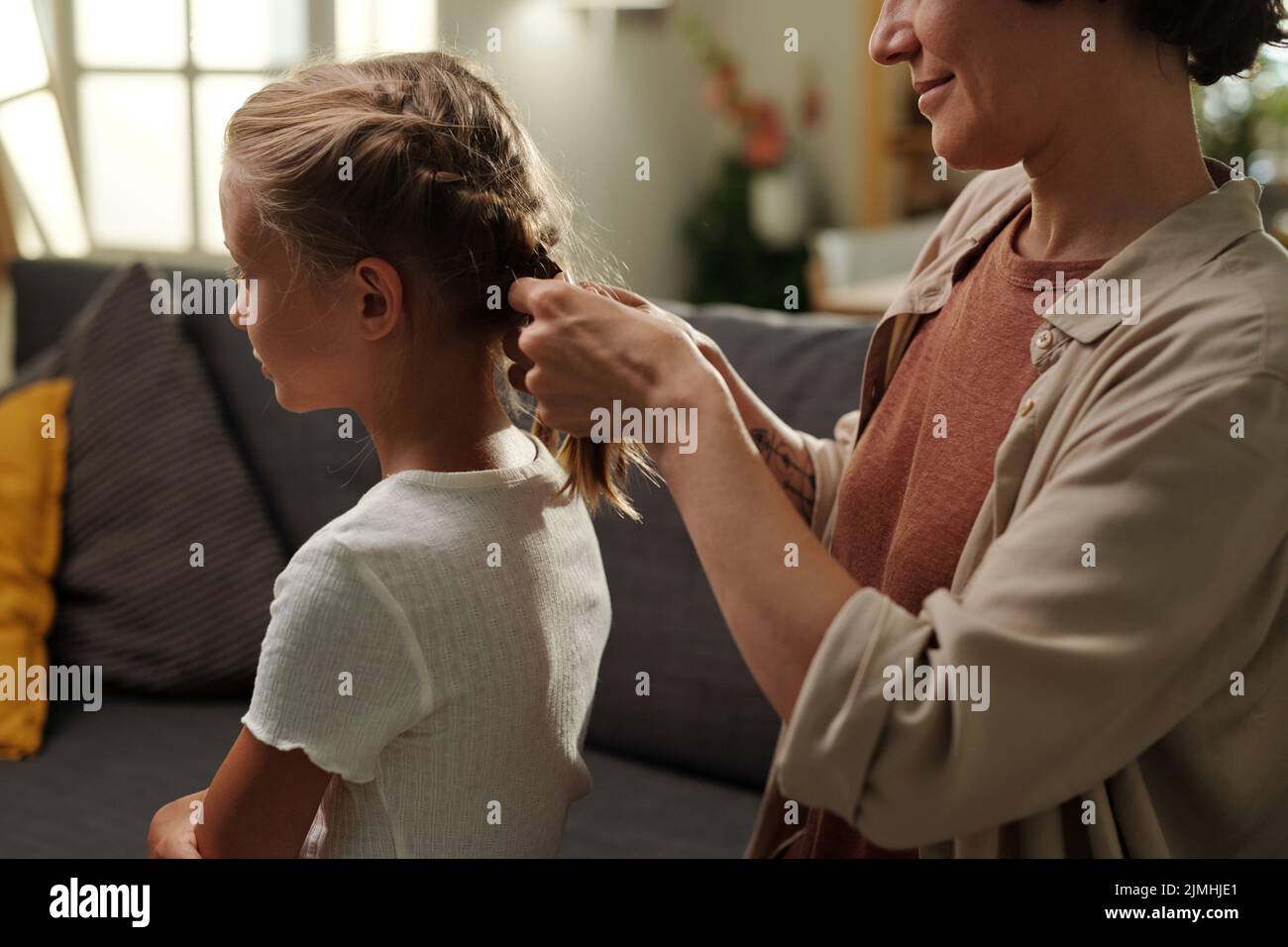 Giovane donna in casualwear intrecciando trecce sulla testa della sua figlia giovane carina con lunghi capelli biondi mentre entrambi seduti sul divano Foto Stock