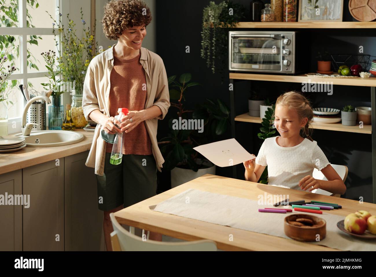 Ragazza felice giovane con carta che mostra il suo disegno alla madre con detergente in bottiglia di plastica che esegue le faccende domestiche Foto Stock