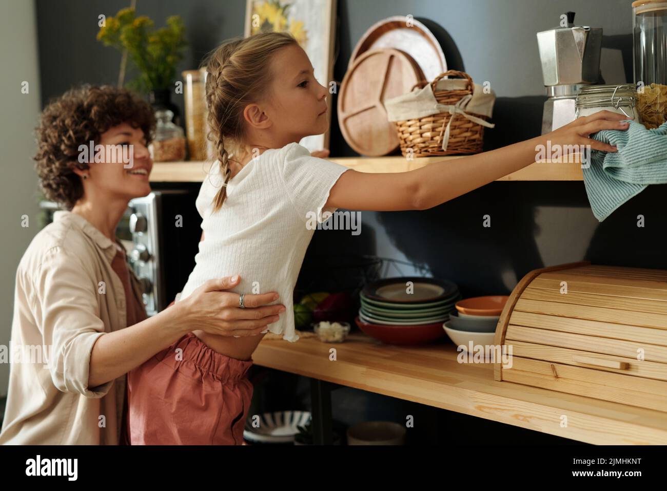 Ragazza giovane carina con spolverare scaffali di legno con utensili da cucina mentre sua madre la sostiene durante le faccende domestiche Foto Stock