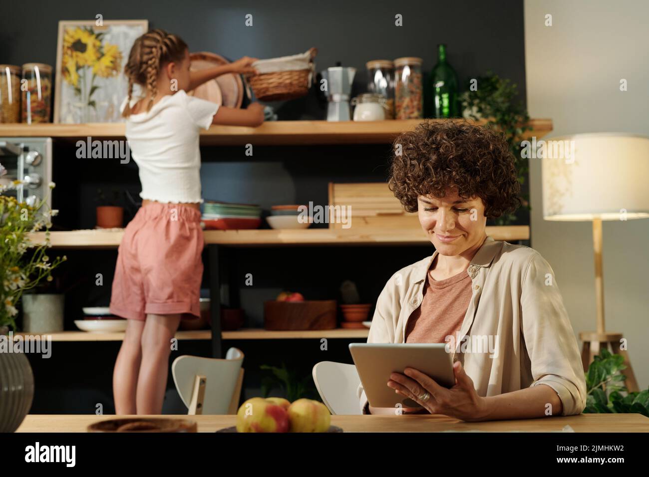 Giovane donna d'affari con la rete del tablet dal tavolo contro la sua figlia giovane che strofinano gli scaffali di legno con gli utensili da cucina Foto Stock