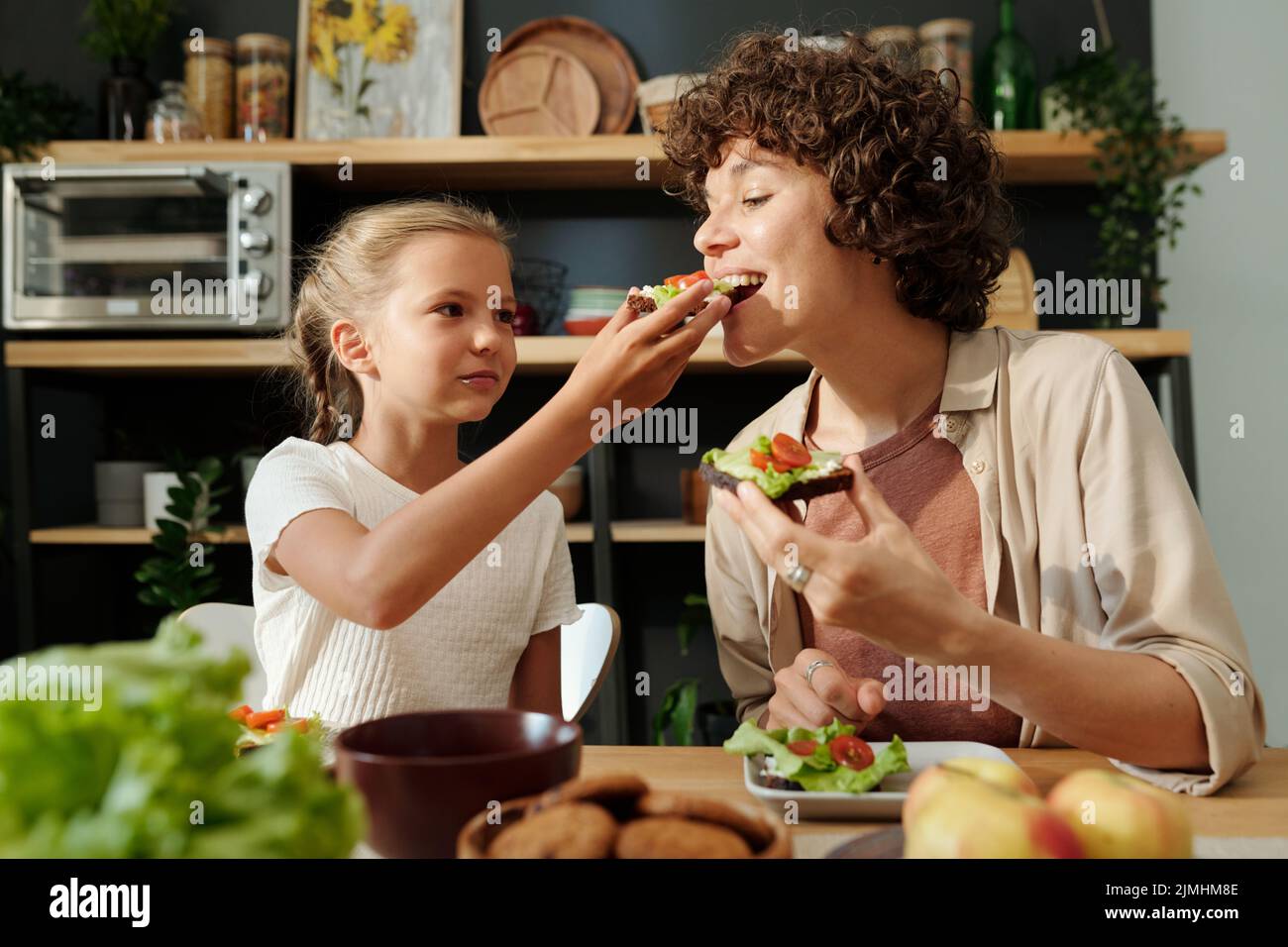 Ragazza giovane carina che dà sua madre appetitoso sandwich per gustare durante la prima colazione, mentre entrambi seduti accanto al tavolo da cucina servito con cibo Foto Stock