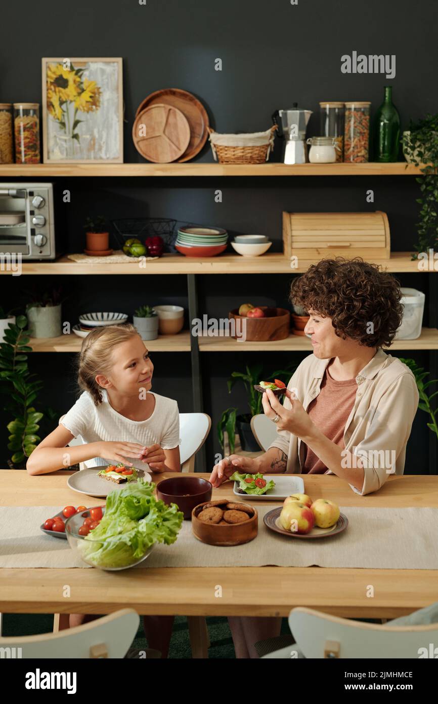 Felice madre giovane e giovane figlia mangiare sandwich per la prima colazione e chiacchierare mentre si siede al tavolo servito Foto Stock