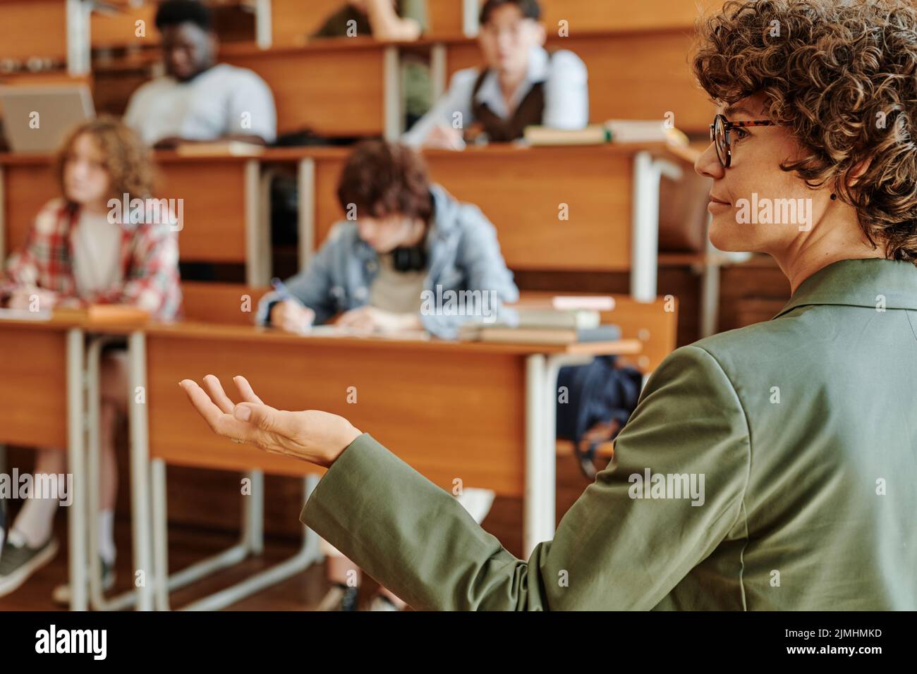 Vista laterale del giovane allenatore fiducioso che spiega il nuovo argomento della lezione al gruppo di studenti multiculturali mentre punta alla lavagna Foto Stock