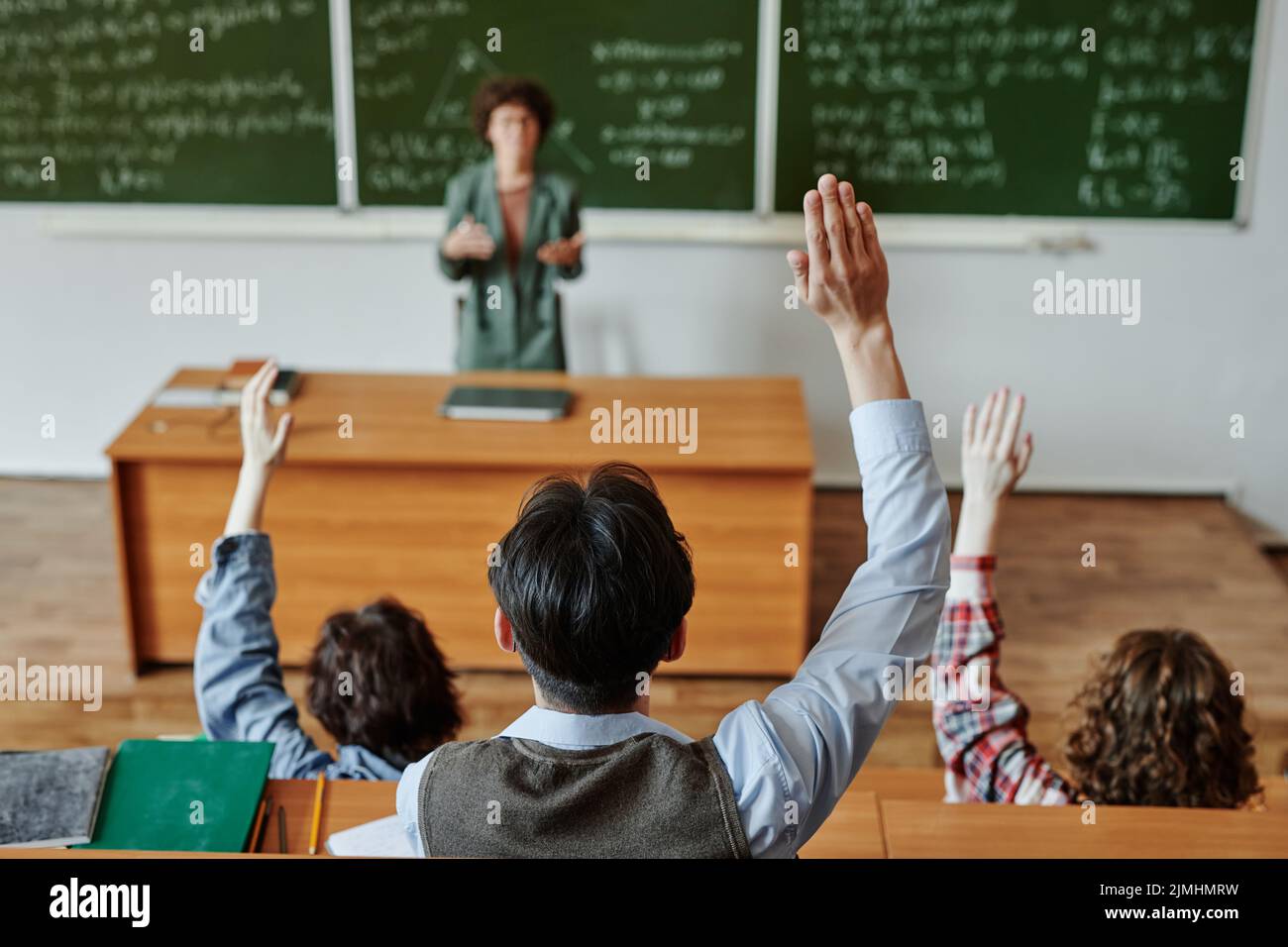 Vista posteriore di diversi studenti giovani tenendo le mani sollevate mentre si siedono da una scrivania di fronte all'insegnante spiegare ne wsubject a lezione Foto Stock