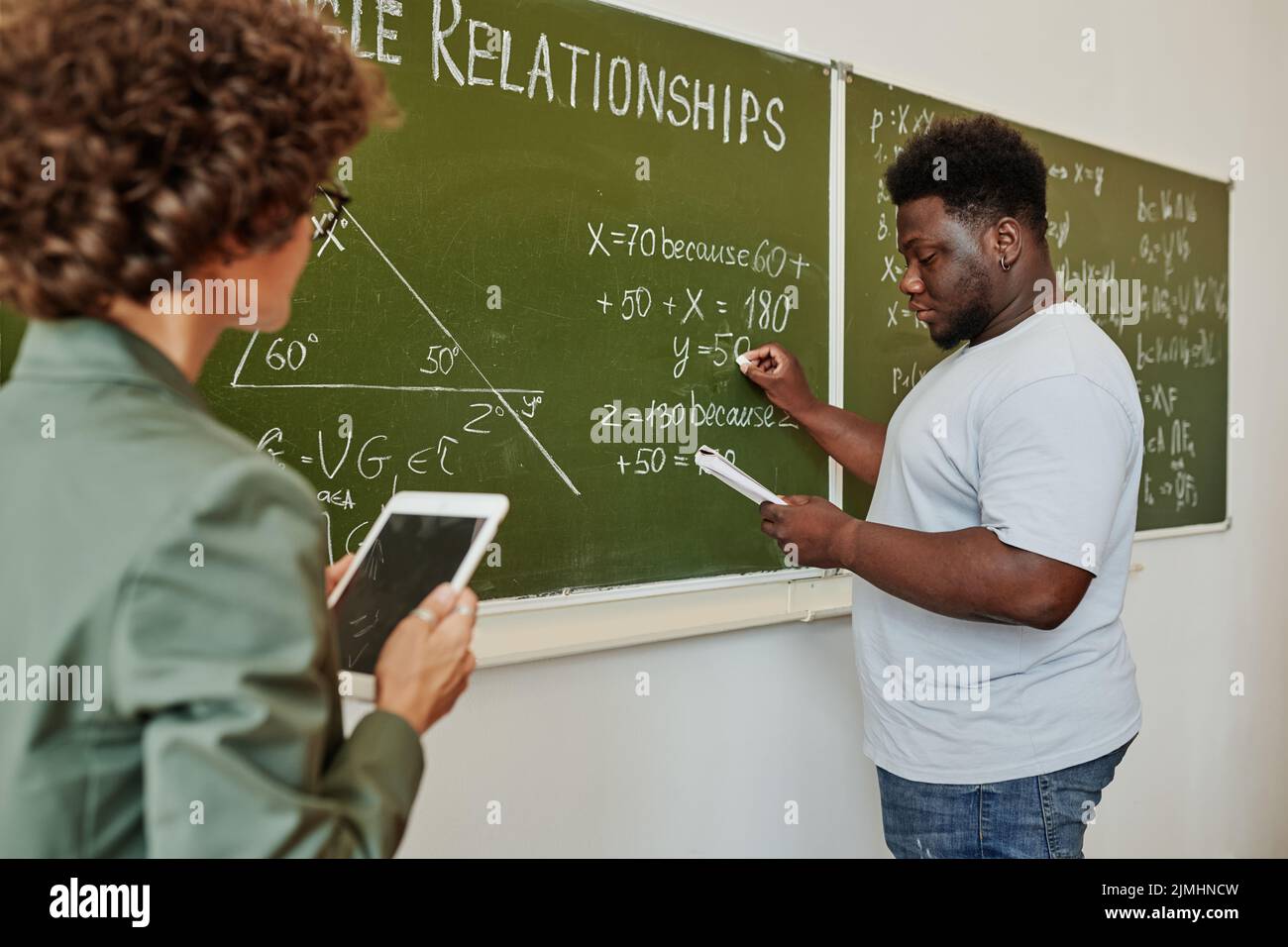 Studente africano americano che scrive formula algebrica o equazione sulla lavagna e che lo spiega all'insegnante a lezione Foto Stock