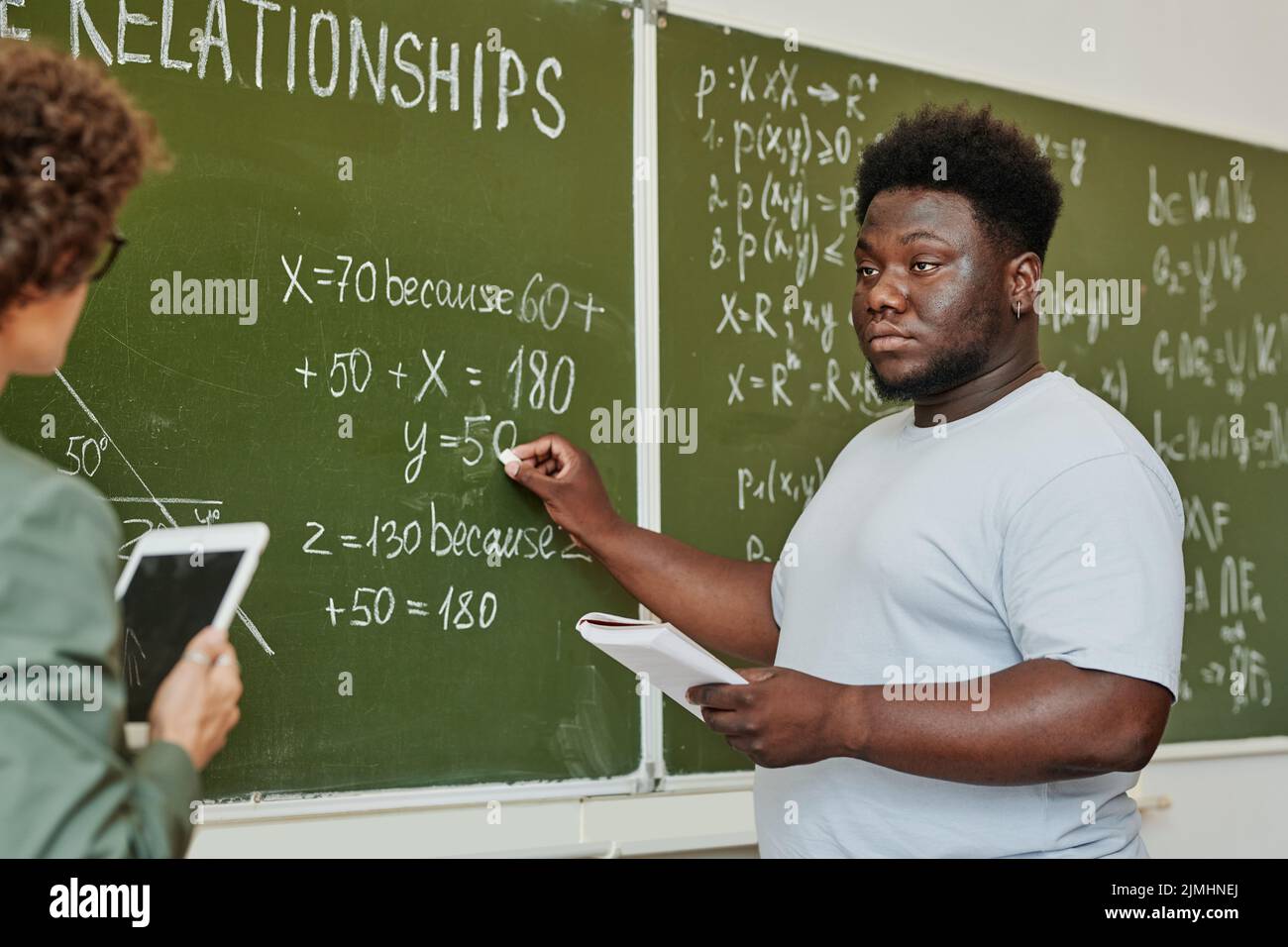Studente afroamericano giovane che punta alla lavagna durante la spiegazione dell'equazione o del teorema e che guarda l'insegnante Foto Stock