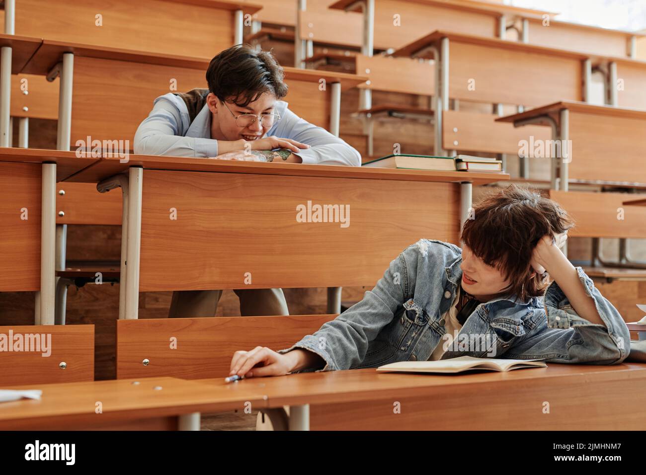 Due giovani studenti interrazziali in casualwear seduti da scrivanie in auditorium a pausa, rilassante e chiacchierando Foto Stock