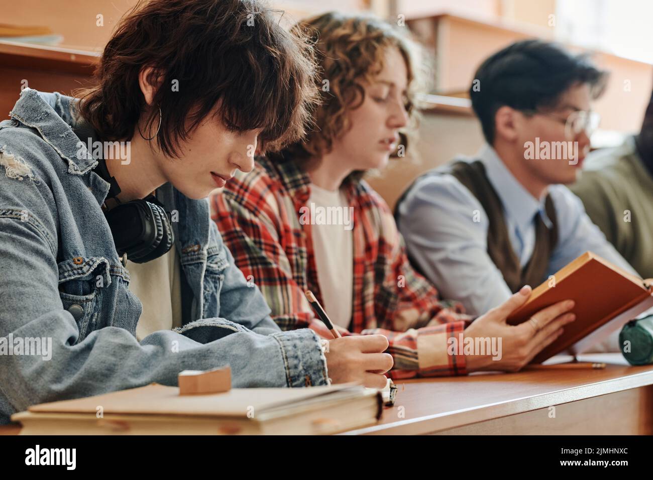 Fila di molti adolescenti interculturali in casualwear che fanno note e libro di lettura mentre si siede dalla scrivania in sala conferenze Foto Stock