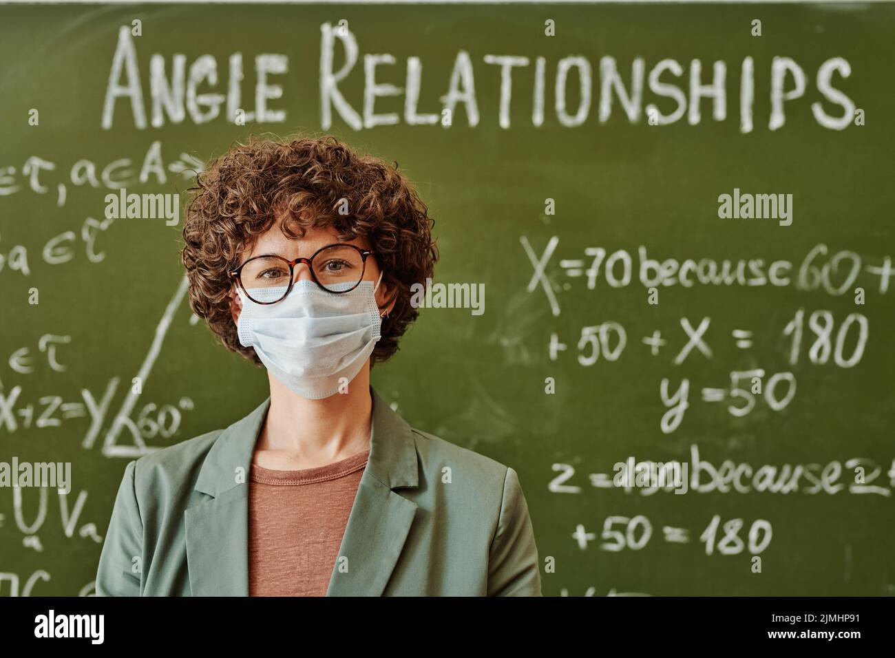 Giovane insegnante di algebra o fisica in occhiali e maschera protettiva in piedi da lavagna con formule ed equazioni Foto Stock