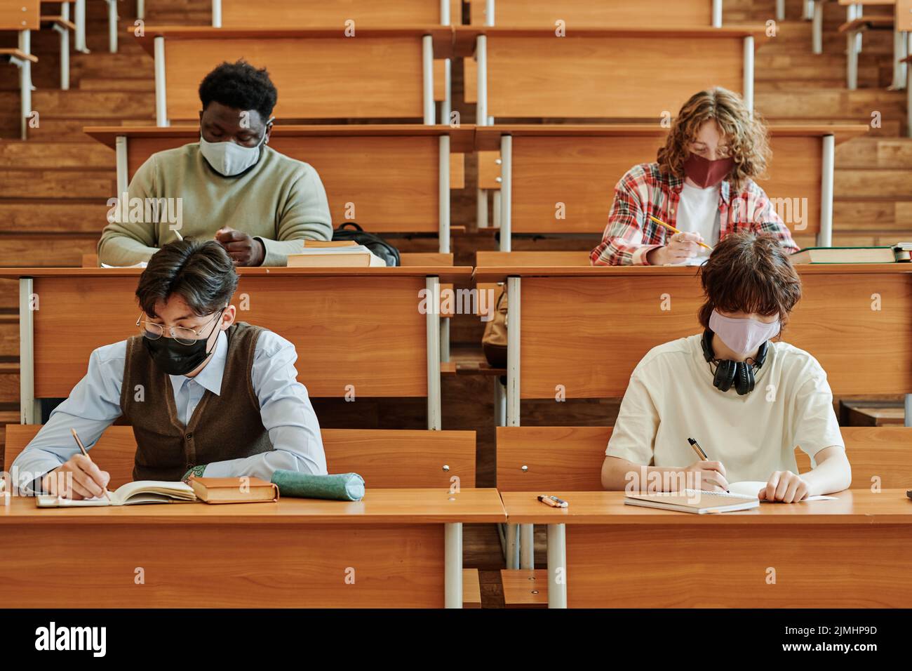 Gruppo di studenti adolescenti in maschere protettive che svolgono compiti individuali mentre si siedono da lunghe scrivanie nella sala conferenze dell'università Foto Stock