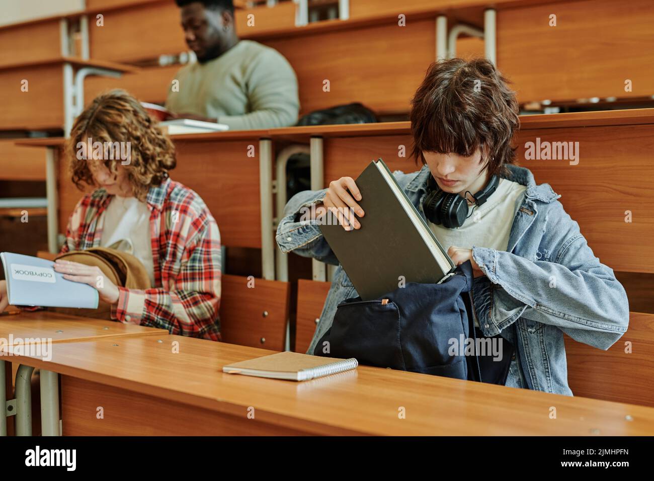 Ragazza bruna adolescente in casualwear mettendo il libro in zaino dopo lezione mentre si siede dalla scrivania in auditorium contro i suoi compagni di classe Foto Stock