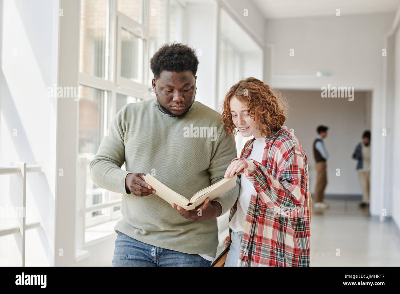 Due studenti di adolescenti interrazziali in casualwear leggendo il libro dalla biblioteca mentre camminano lungo il corridoio dell'università prima della lezione Foto Stock