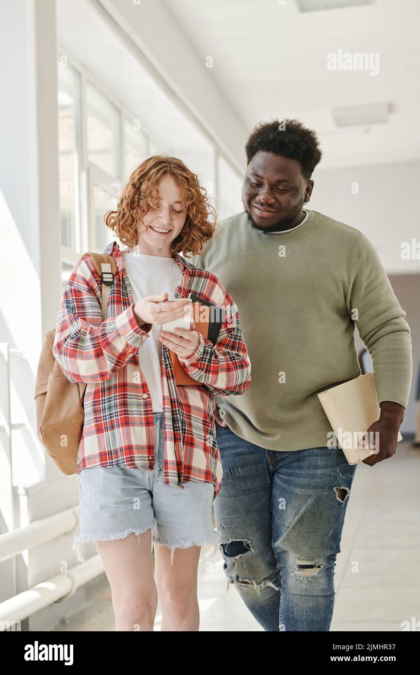 Due compagni di classe adolescenti in casualwear che parlano di curiosità in smartphone tenuto da ragazza sorridente carino dopo le lezioni Foto Stock