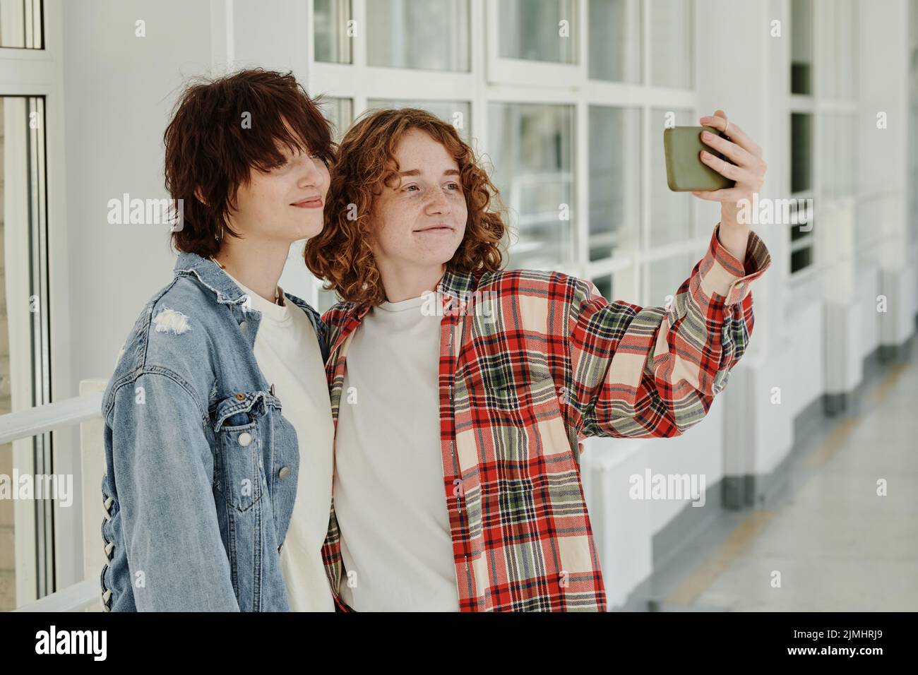 Due ragazze adolescenti felici che prendono selfie nel corridoio del college alla pausa mentre si levano in piedi l'una vicino all'altra e guardano la fotocamera dello smartphone Foto Stock