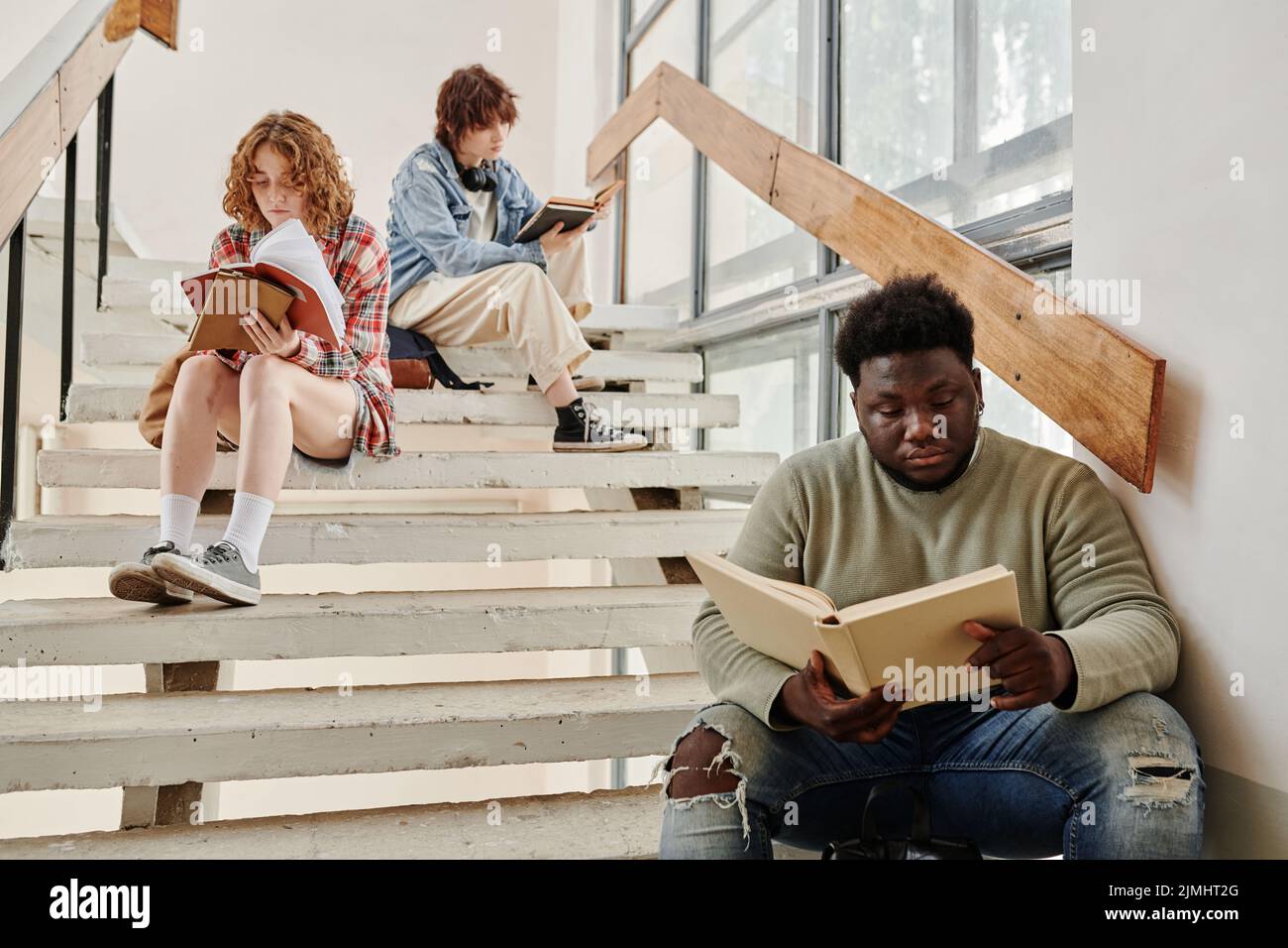 Giovane studentessa afroamericana e le sue due compagni di classe femminili leggendo libri mentre si siede su scala nel corridoio dell'università alla pausa Foto Stock