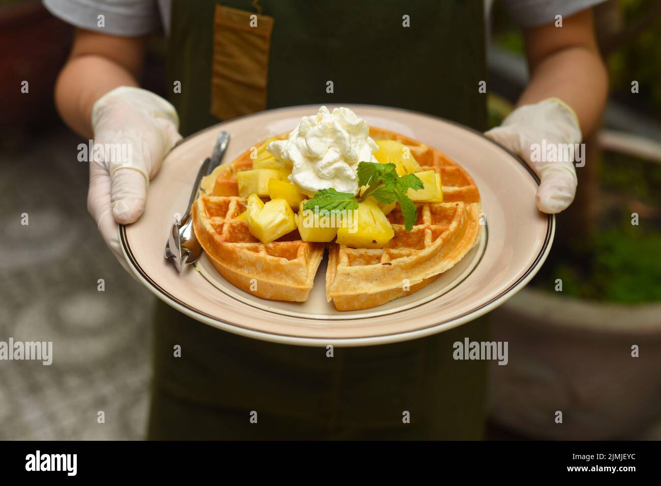 Mani in guanti di gomma che tengono waffle belgi con ananas e gelato su un piatto Foto Stock