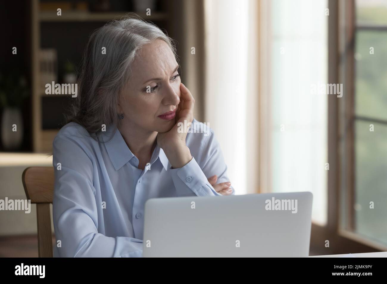 Donna anziana seria e premurosa seduta al computer portatile a casa Foto Stock