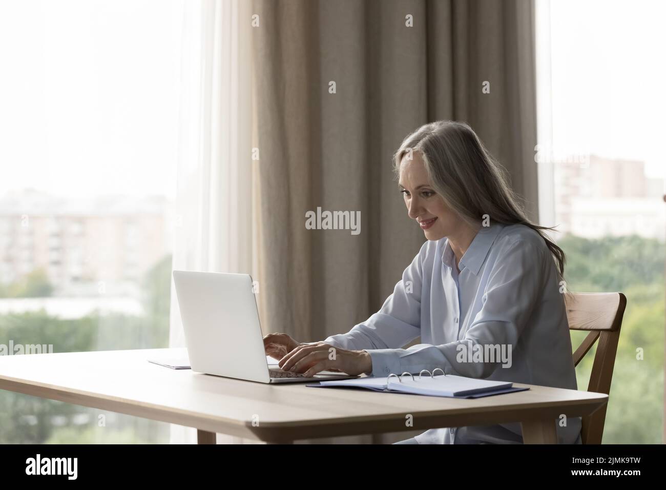 Donna anziana positiva focalizzata che usa il computer portatile in ambienti chiusi Foto Stock
