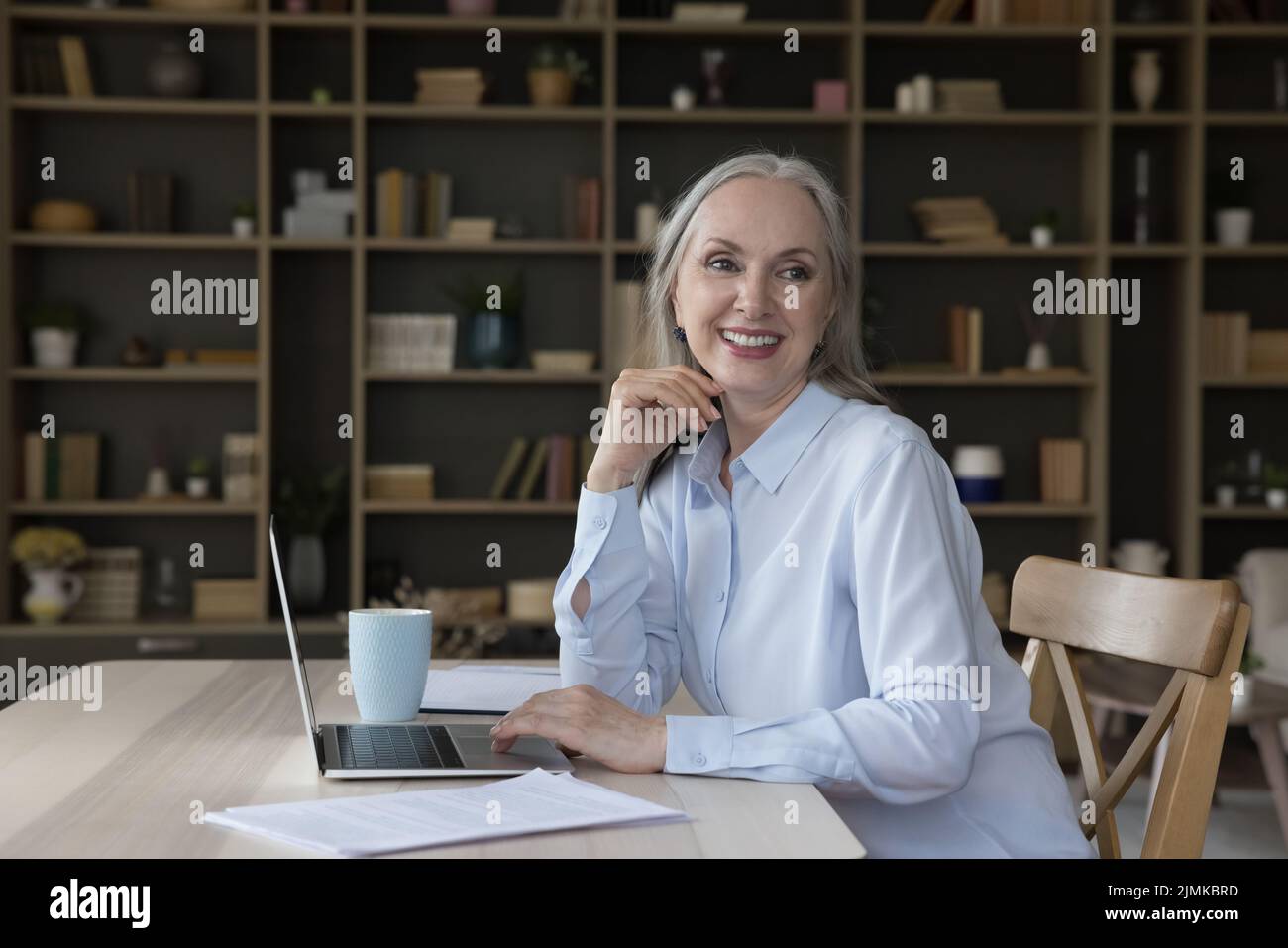 Gioioso felice bella anziana donna d'affari freelance che guarda lontano dal tavolo Foto Stock