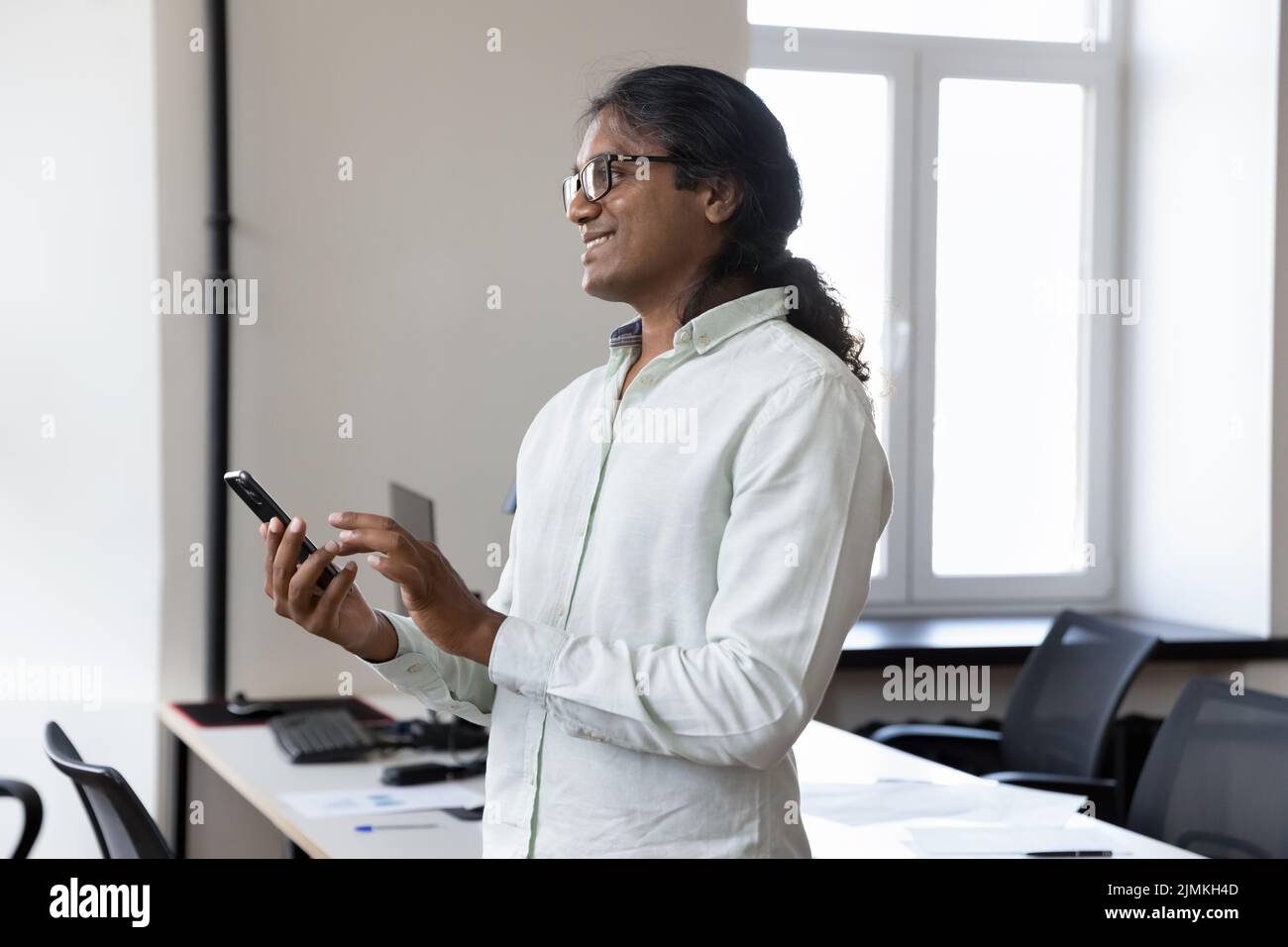 Pensieroso uomo imprenditore indiano in piedi in ufficio con smartphone Foto Stock