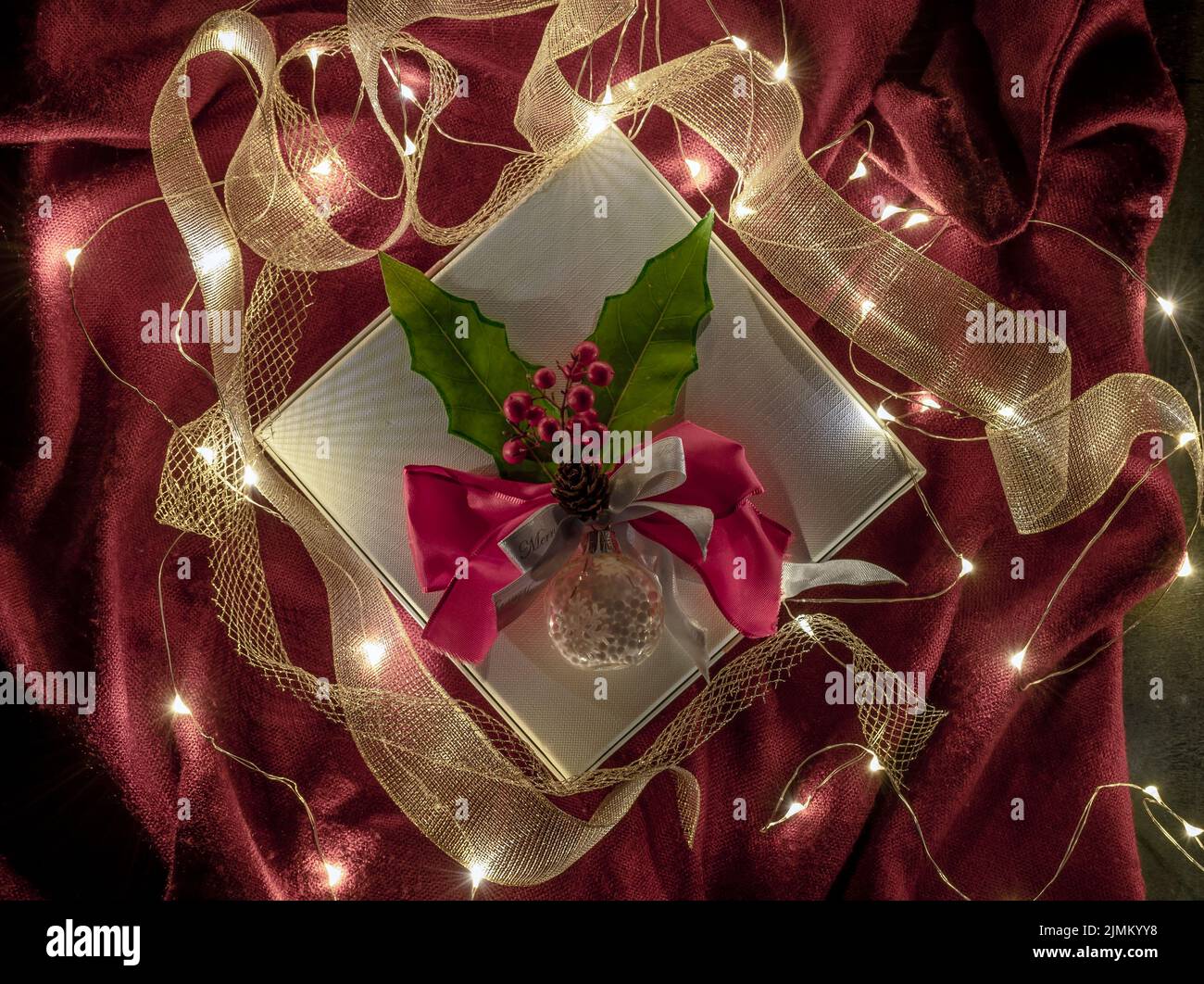 Un regalo di Natale è decorato con nastri d'oro, verde e rosso agrifoglio e luci fiabe incandescenti su uno sfondo rosso Foto Stock