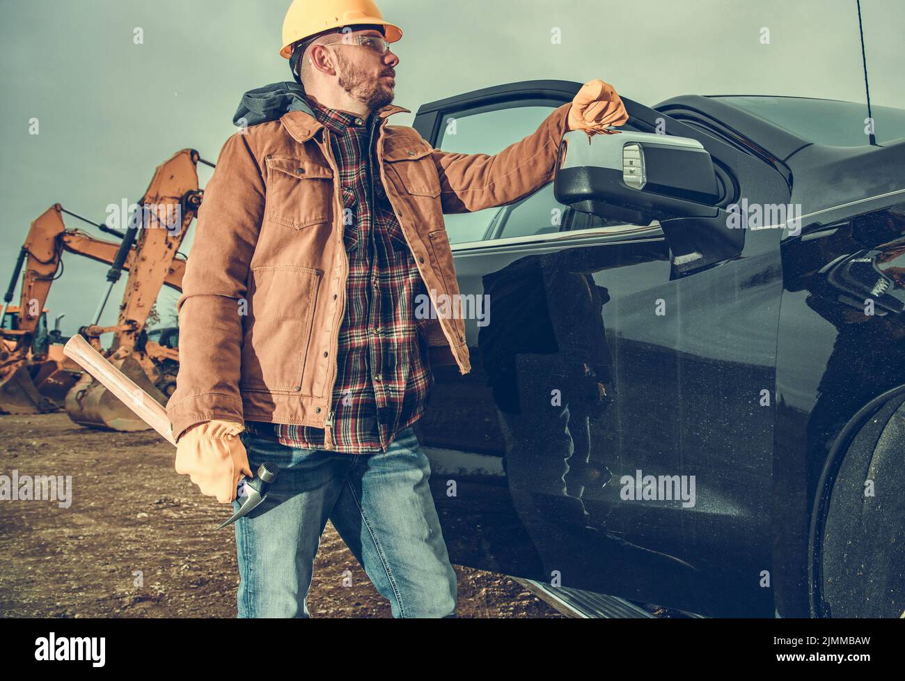 Orgoglioso lavoratore edile per impieghi gravosi in piedi accanto al pick-up nero con escavatori in background. Sensazione di soddisfazione dopo aver fatto bene ha Foto Stock