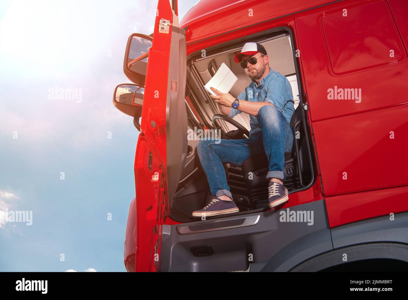 Autista di camion caucasico seduto nella cabina del suo semi-camion rosso che verifica attentamente i documenti di transito prima di iniziare un percorso con un grande carico consegnare Foto Stock