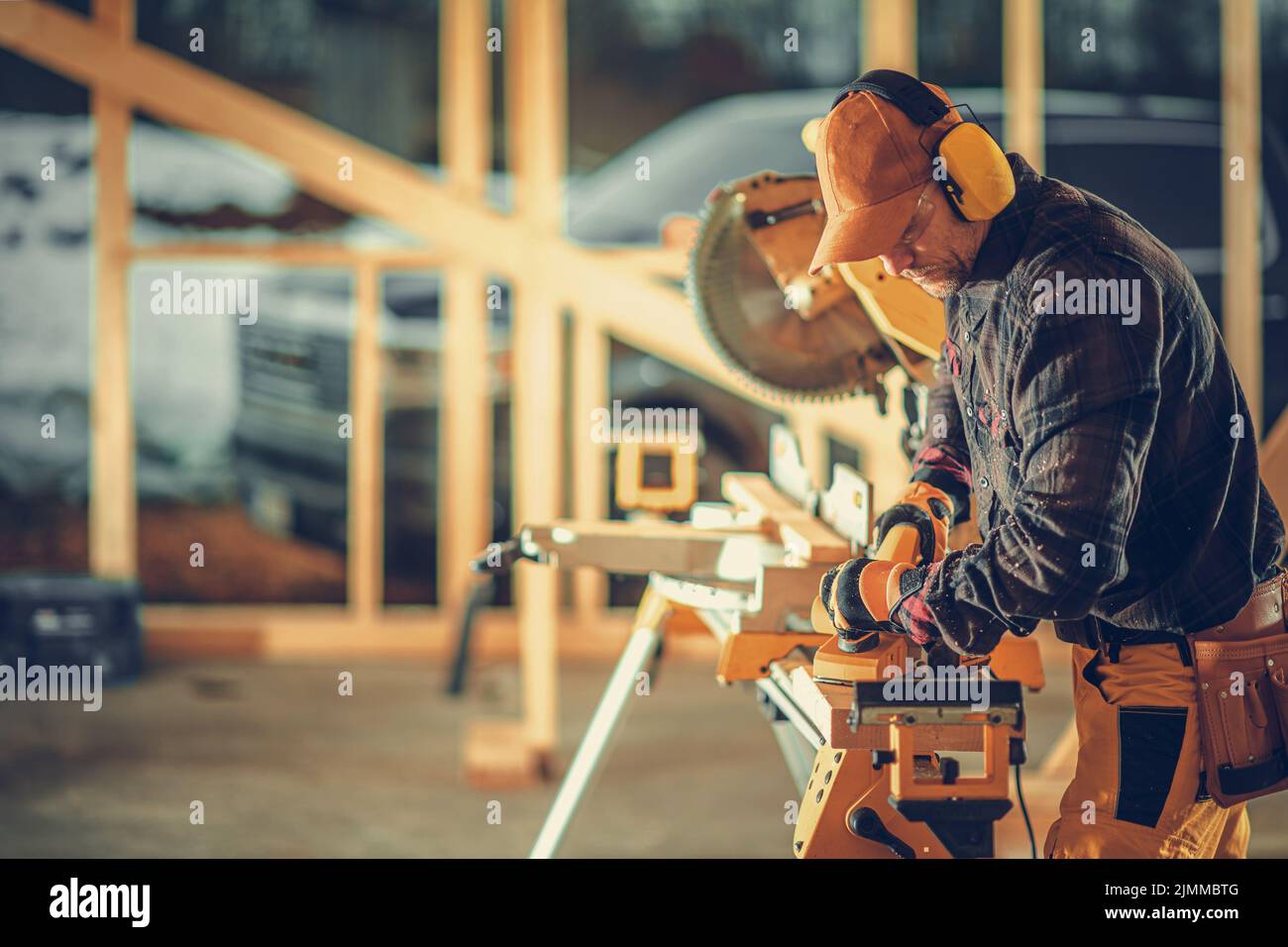 Appaltatore caucasico che lavora con il legno mentre costruisce la cornice di legno di edificio residenziale di stile canadese. Cantiere sullo sfondo Foto Stock