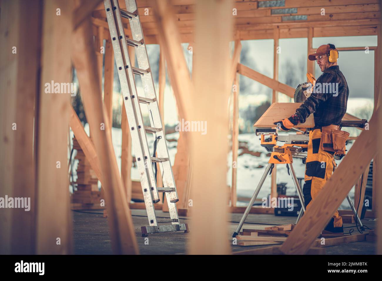 Appaltatore che elabora i pannelli di legno usando il suo Carpentry Equipment. Casa di costruzione in stile canadese sito in background. Industriali Foto Stock