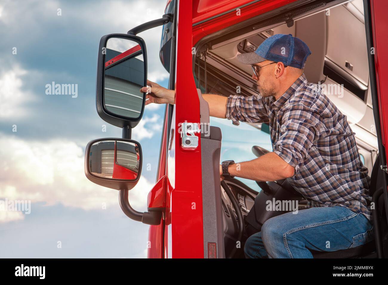 Maschio caucasico Trucker seduto all'interno della cabina del suo camion rosso adattando lo specchio di vista laterale. Preparazione prima di una guida lunga. Trasporto per impieghi gravosi Foto Stock