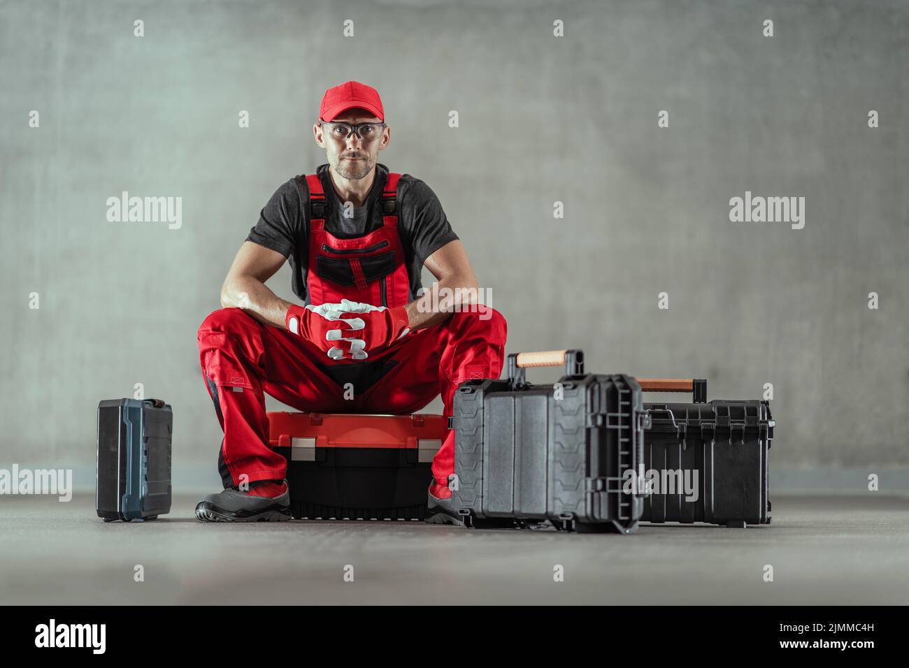 Handyman caucasico in Red Work uniforme seduto sul Toolbox circondato da più scatole dei suoi attrezzi di riparazione professionale. Sfondo grigio solido. Horizo Foto Stock