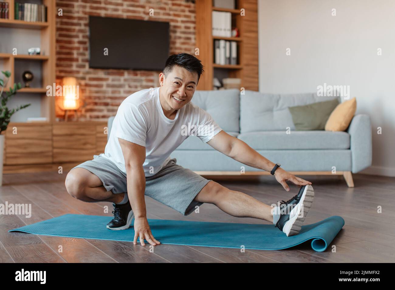 Felice asiatico uomo maturo formazione a casa, allungando le gambe in soggiorno interno, godendo di stile di vita sano Foto Stock